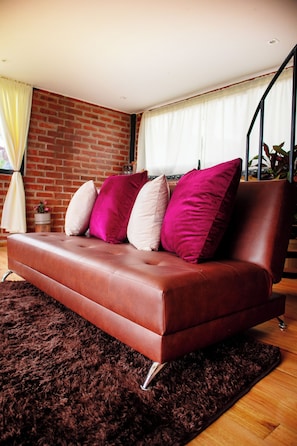 Ein braunes Ledersofa mit vielen Kissen in einem Raum, der eine Ziegelwand, Holzboden und ein großes Fenster mit Vorhängen zeigt.