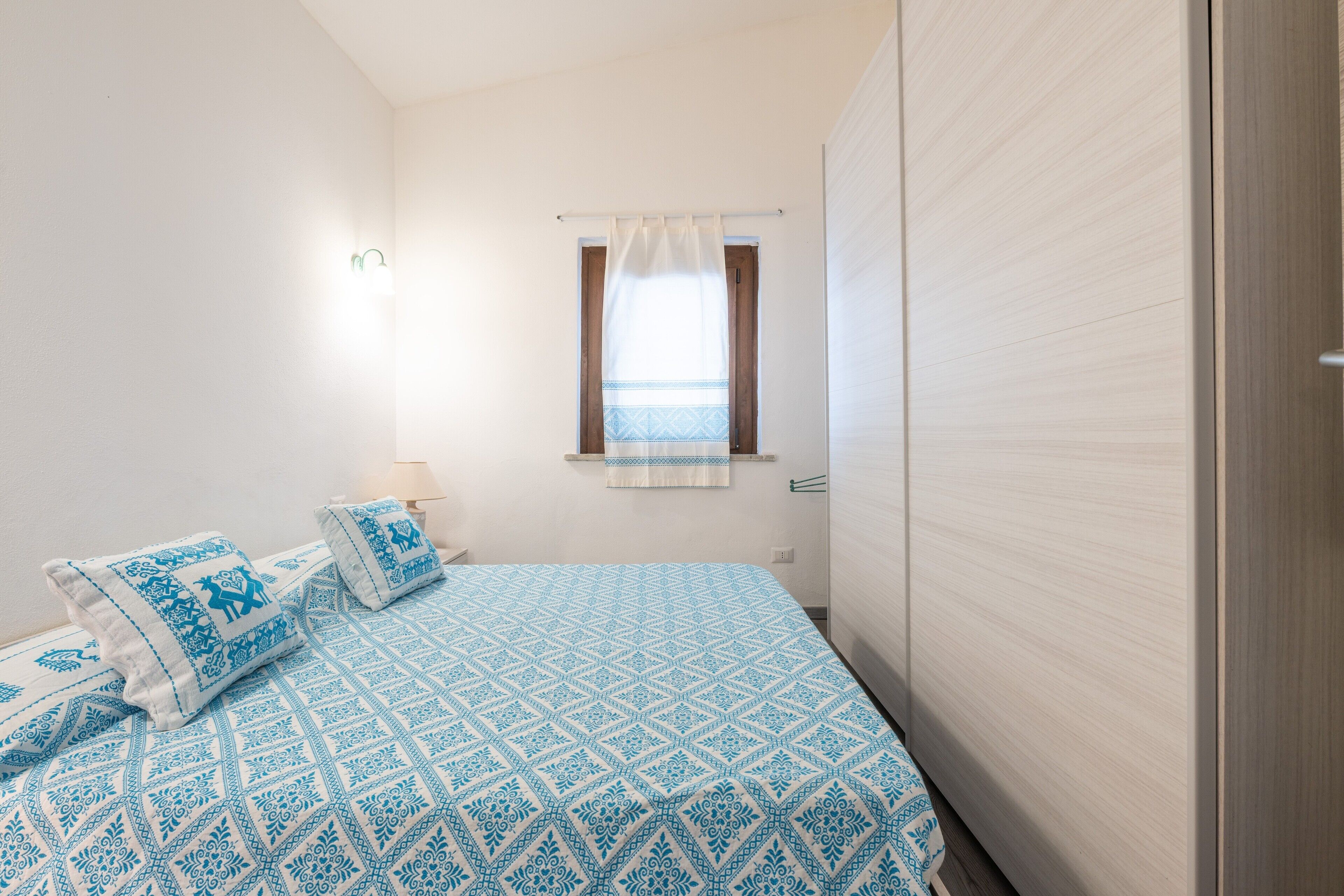 A bedroom with a bed, pillows, a window with curtains, a wall-mounted light, and a wooden wardrobe.