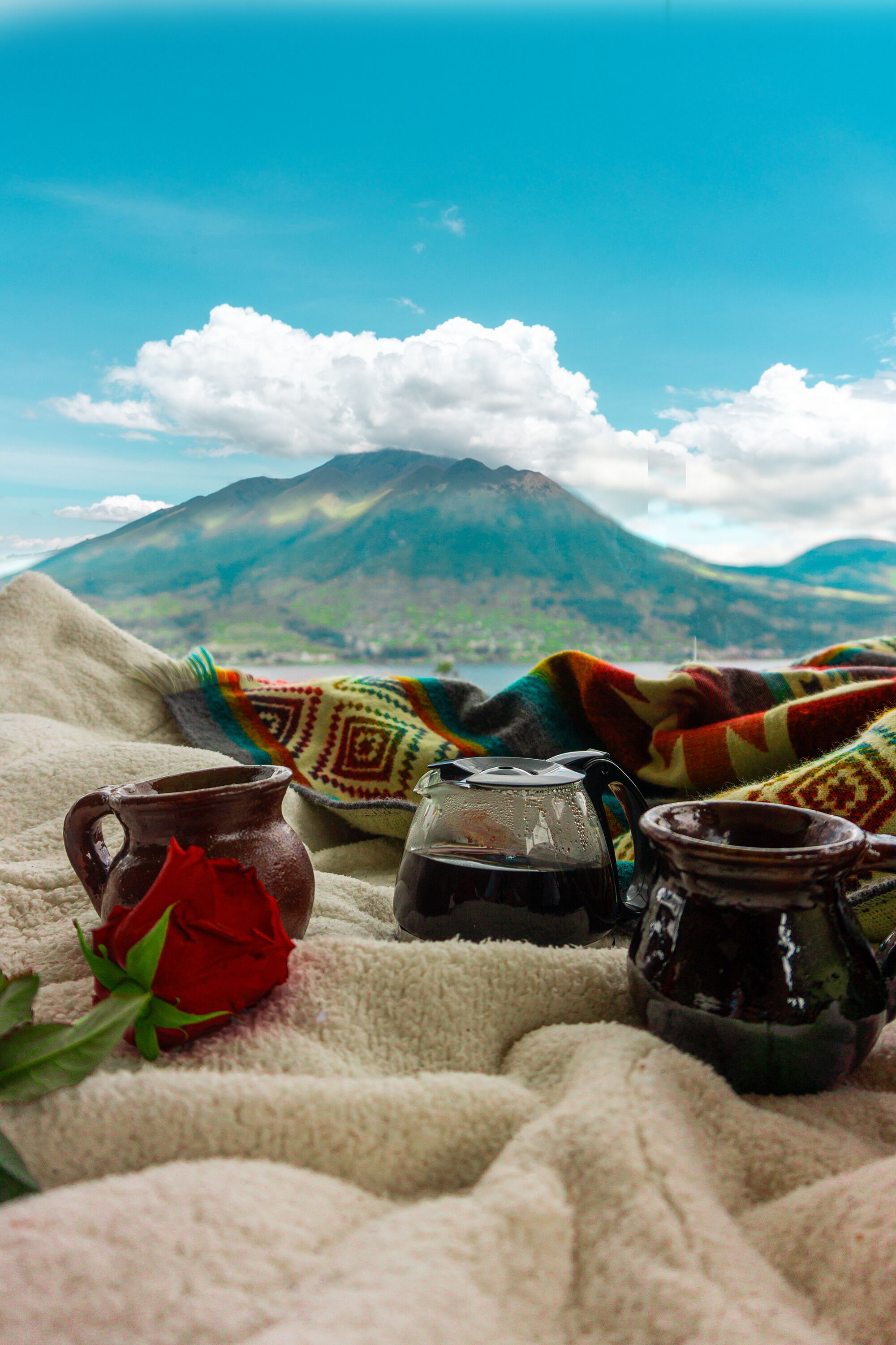 A cozy blanket with a colorful pattern, a red rose, and two small ceramic mugs on a soft surface.