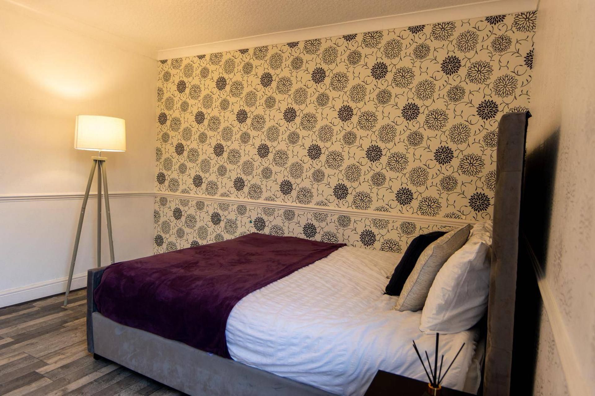 A hotel room with a bed, a floor lamp, and a patterned wallpaper.