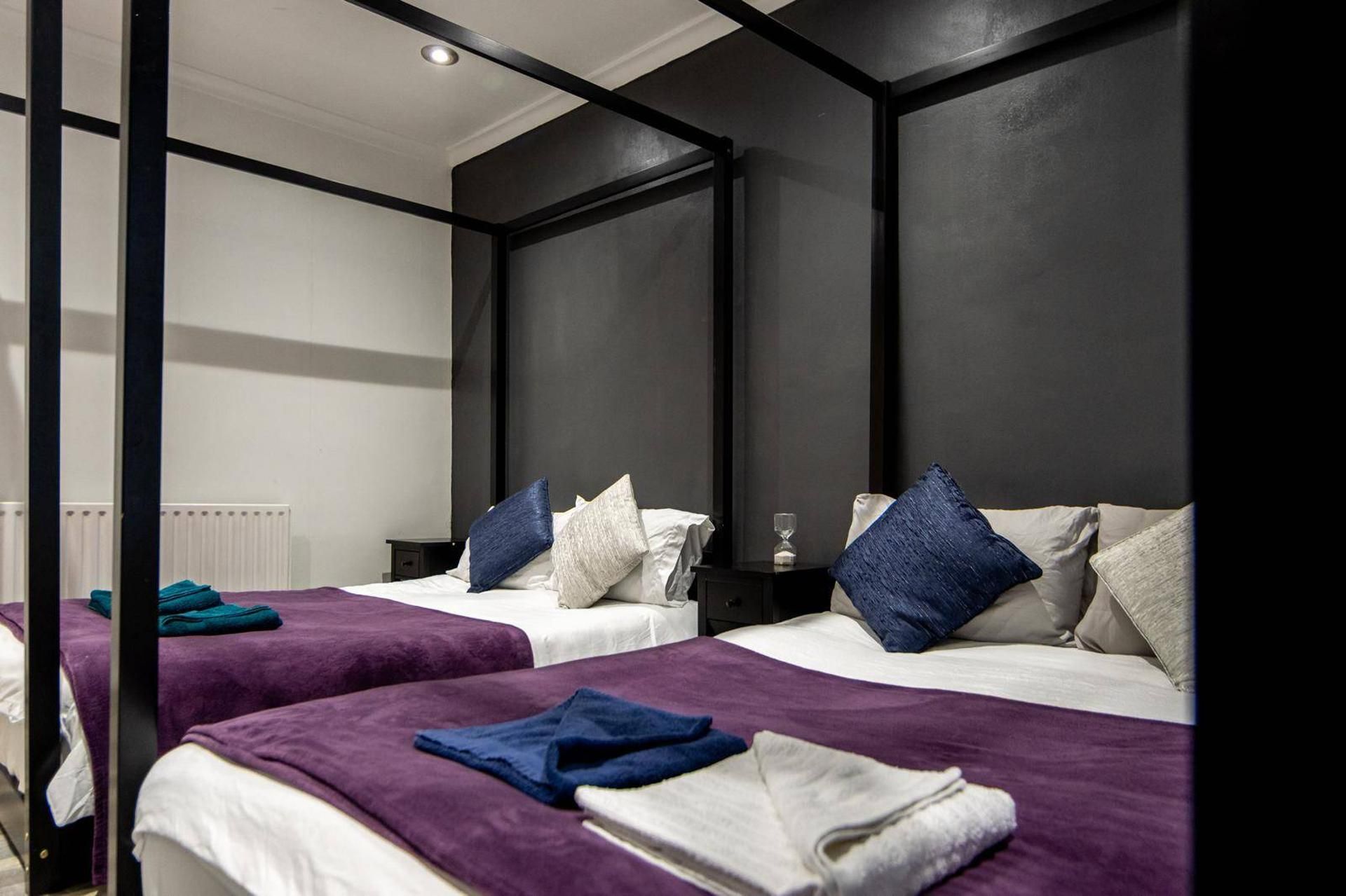 A hotel room with two beds, each with a purple bedspread and white linens. There is a small bedside table with a glass and a book. The room has dark walls and a ceiling light.