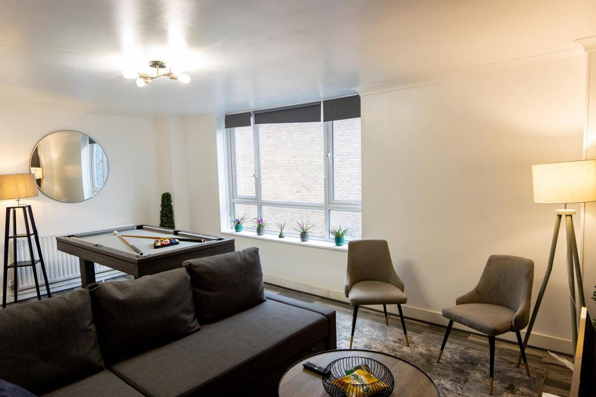 Una sala de estar moderna con una mesa de billar, un sofá, dos sillones, un espejo redondo y una ventana con vistas a un edificio de ladrillo.