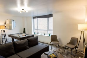 Una sala de estar moderna con una mesa de billar, un sofá, dos sillones, un espejo redondo y una ventana con vistas a un edificio de ladrillo.
