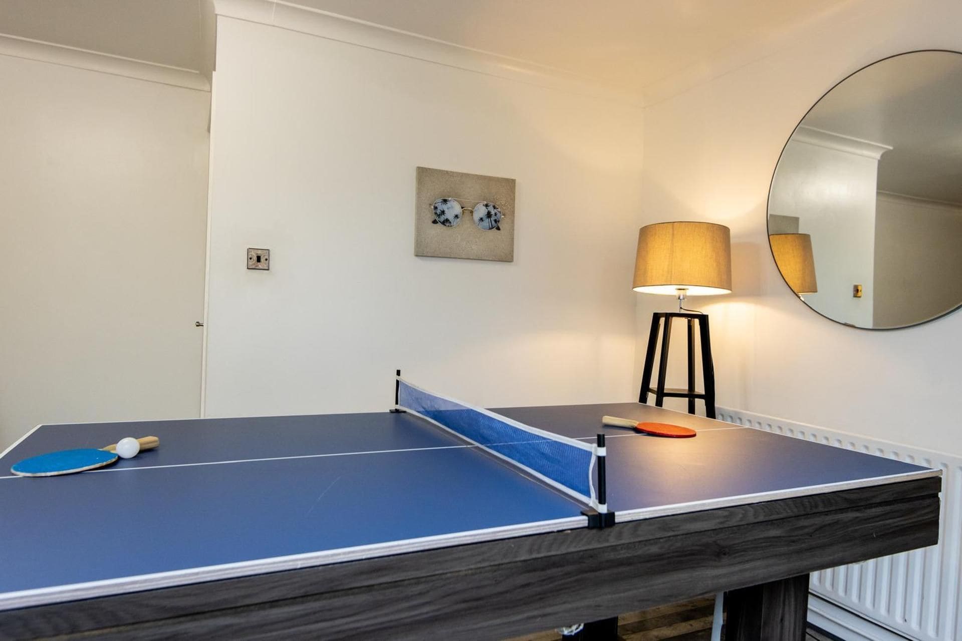 A ping pong table with a blue playing surface and a net in a room with white walls, a round mirror, and a lamp.