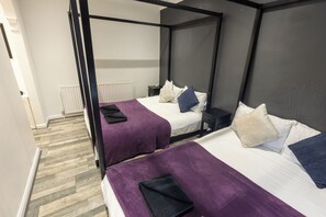 A four-poster bed room with two beds, each with a purple bedspread and pillows, and a white radiator in the background. - Housesleep14-freep-garden-bbq-patio-pooltable (London)
