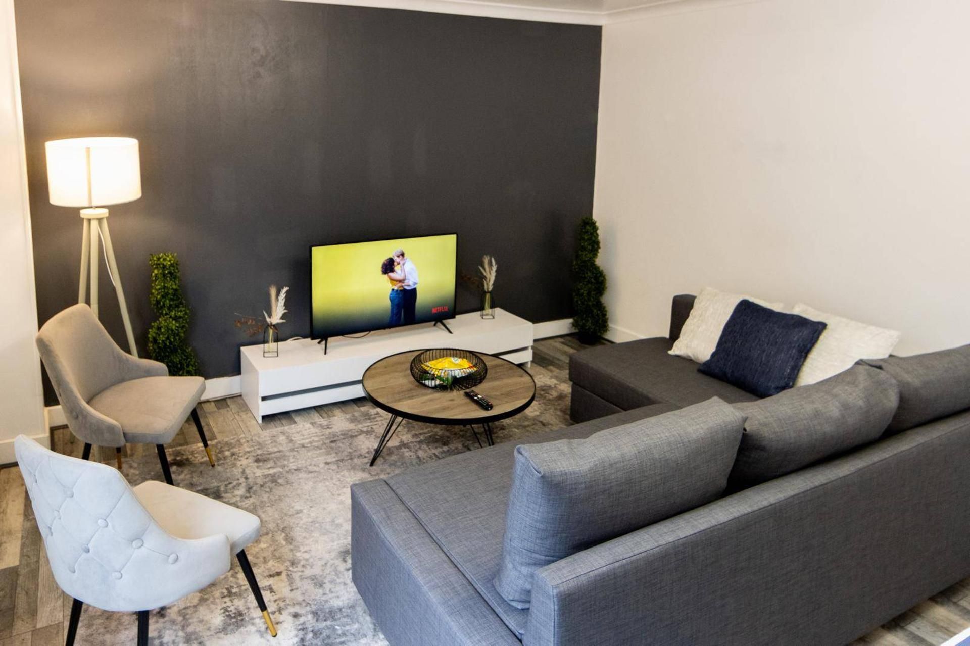 A modern living room with a grey sofa, a white armchair, a round coffee table, and a flat-screen TV on a white stand.
