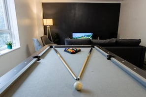 Ein Billardtisch mit Spielbällen und einer Billardkugel, ein Fernseher im Hintergrund und ein Sofa.