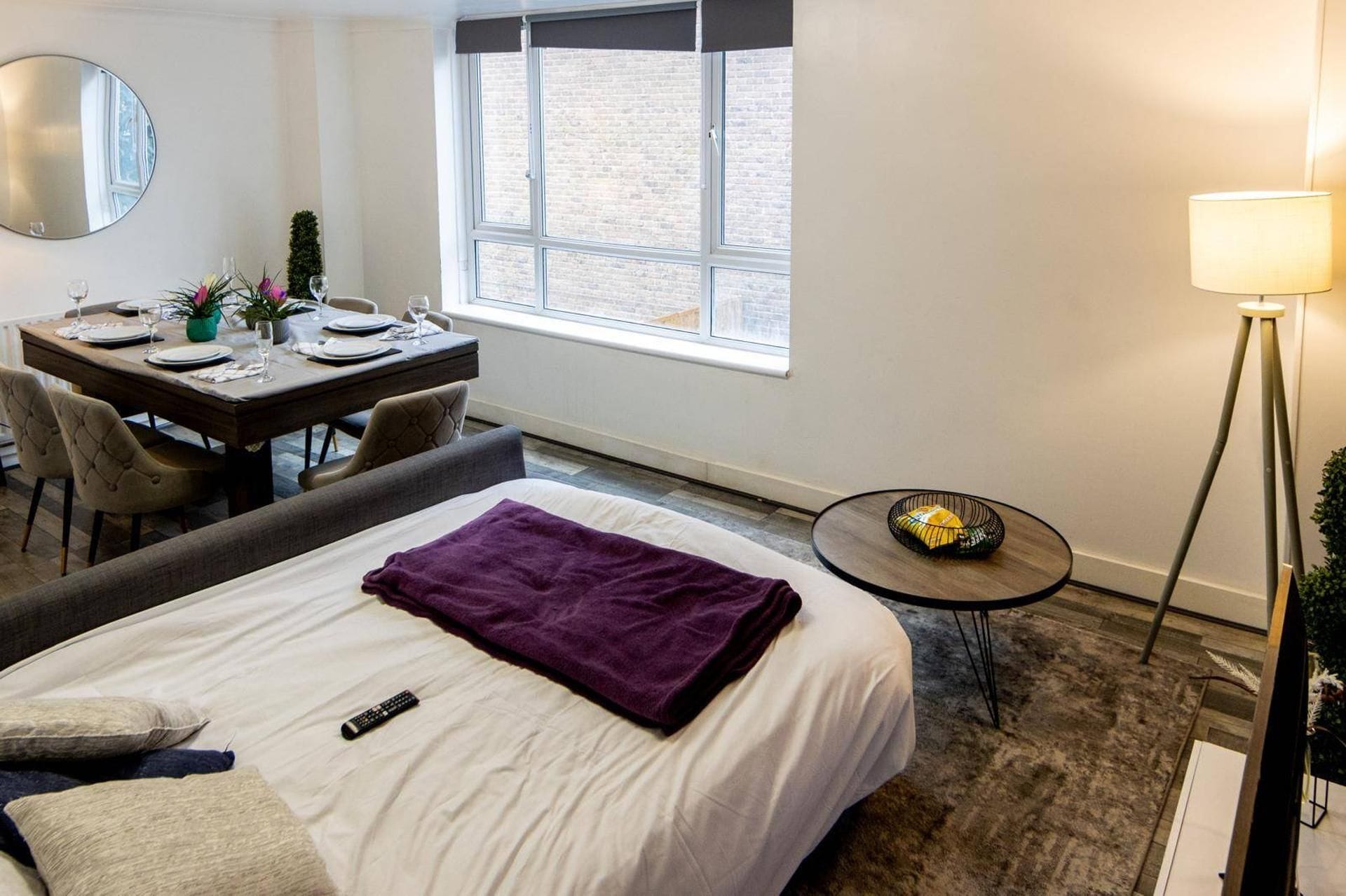 Habitación de hotel con cama, escritorio con silla, una mesita, una lámpara y ventana con vista a un edificio de ladrillo.