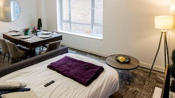 Habitación de hotel con cama, escritorio con silla, una mesita, una lámpara y ventana con vista a un edificio de ladrillo.