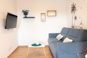 A living room with a grey sofa, a small round table with a green coaster, a flat-screen TV mounted on the wall, and a wall decoration of palm trees and birds.