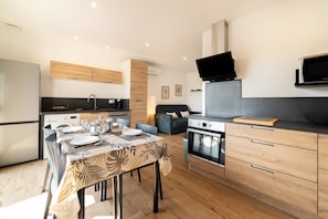 A modern kitchen with a dining table set for four, a built-in oven, and a flat-screen TV mounted above the counter. - Charming 'Côté Nature' Cottage with Mountain View, Wi-Fi & Air Conditioning (Sorède)