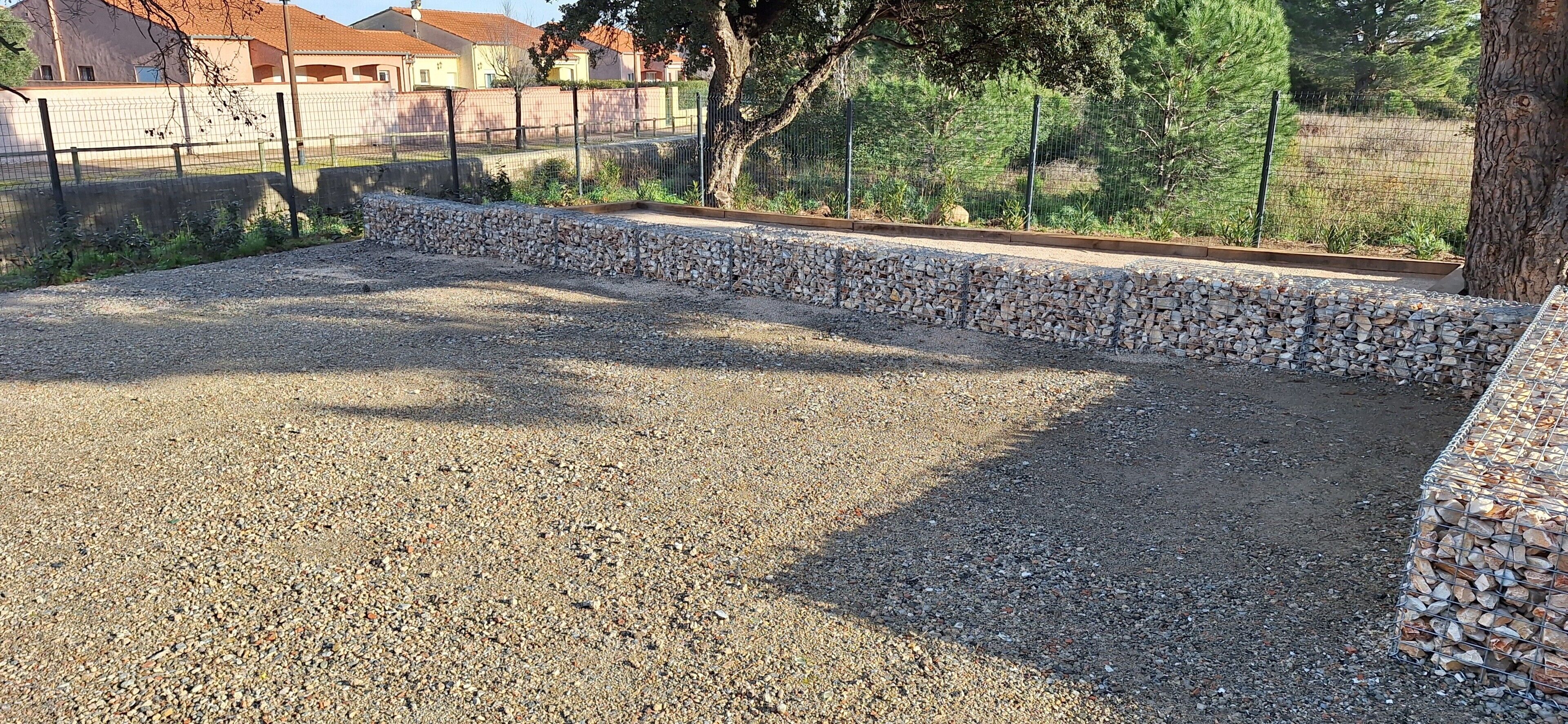 Um pátio de cascalho com uma borda de pedra, uma árvore e uma cerca ao fundo.