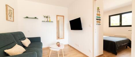 Ett rum med en grön soffa, ett litet vitt bord, en tv som är monterad på väggen, en säng med ett svart överkast och ett fönster med persienner.