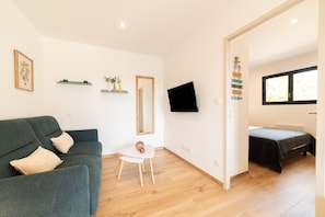 A room with a green sofa, a small white table, a TV mounted on the wall, a bed with a black bedspread, and a window with blinds. - Charming 'Côté Nature' Cottage with Mountain View, Wi-Fi & Air Conditioning (Sorède)