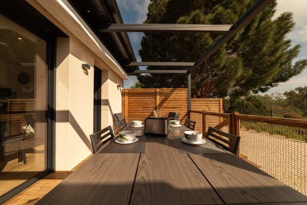 An outdoor patio with a wooden table set for dining, a toaster, and a coffee maker.