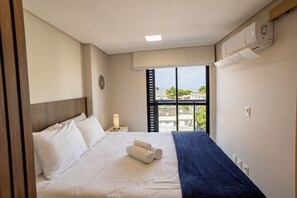 A hotel room with a bed, a nightstand, a lamp, a window with blinds, and an air conditioning unit.