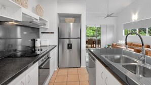 A modern kitchen with stainless steel appliances, a black countertop, and a view to a dining area. - Wallaby Outlook- Tranquil Trinity Beach Retreat | Pool, Balcony & Nature Views (Trinity Beach)
