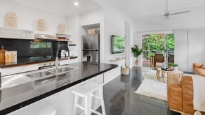 A modern kitchen with a central island, stainless steel appliances, and a dining area with a view of greenery. - Wallaby Outlook- Tranquil Trinity Beach Retreat | Pool, Balcony & Nature Views (Trinity Beach)