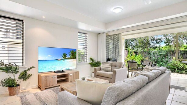A modern living room with a large flat-screen TV, a grey sofa, a coffee table, and a dining area with a table and chairs. - Stylish Cairns Escape with Courtyard | Cosmo 14 (Cairns North)