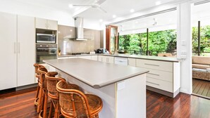 A modern kitchen with a central island, bar stools, and a view of lush greenery. - Spacious 7-Bedroom Queenslander Retreat | Privacy, Comfort & Bushland Serenity (Aeroglen)