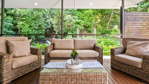 A wicker sofa set with cushions, a glass coffee table with a plant, and a view of lush greenery through a balcony. - Spacious 7-Bedroom Queenslander Retreat | Privacy, Comfort & Bushland Serenity (Aeroglen)