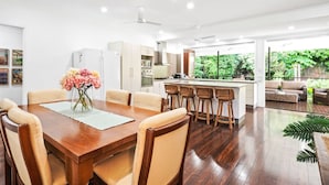 A modern kitchen with white cabinets, a central island with bar stools, and a dining area with a wooden table and chairs. - Spacious 7-Bedroom Queenslander Retreat | Privacy, Comfort & Bushland Serenity (Aeroglen)
