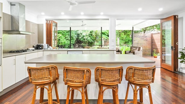 A modern kitchen with a large island, three wicker bar stools, and a view of a lush garden through large windows. - Spacious 7-Bedroom Queenslander Retreat | Privacy, Comfort & Bushland Serenity (Aeroglen)