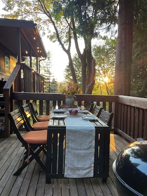 Un espace de restauration en plein air doté d’une terrasse en bois, d’une table dressée avec des assiettes et un centre de table, ainsi que de chaises avec des coussins.