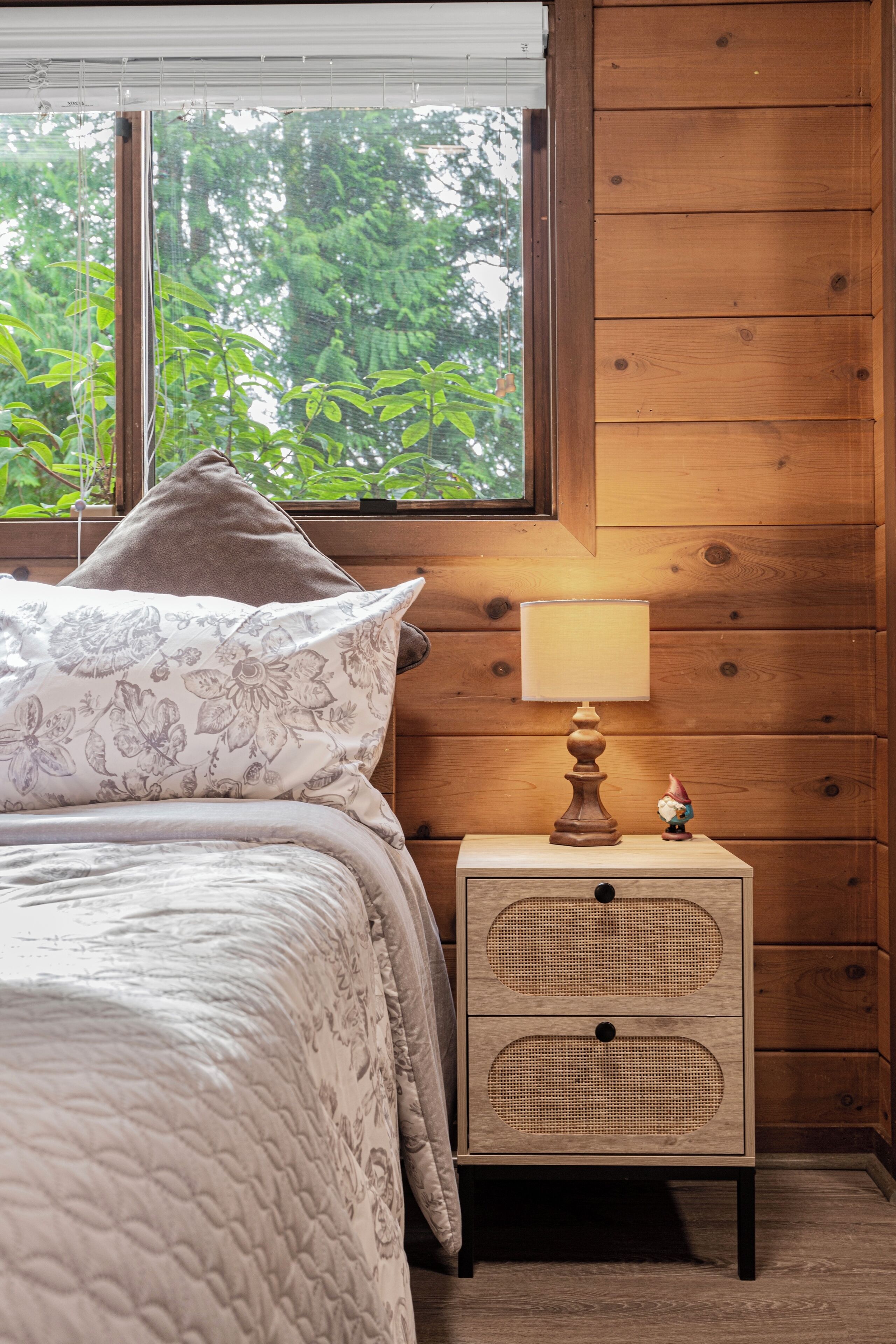 A bedroom with a bed, a nightstand with a lamp, and a window with a view of greenery.