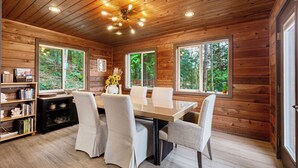 L’intérieur d’un chalet en bois comprend une table à manger, des chaises, des étagères et de grandes fenêtres donnant sur la forêt.