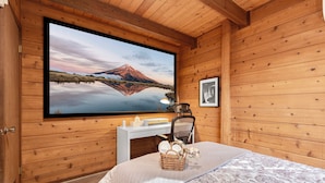 Une chambre de style rustique, avec des murs en bois, un grand téléviseur fixé au mur, un lit recouvert d’une courtepointe à motifs floraux, un panier rempli de serviettes et un tableau encadré accroché au mur.