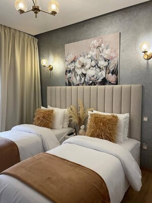 A bedroom with two beds, a grey headboard, a floral painting, and wall sconces.