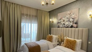 Une chambre d’hôtel avec deux lits, une grande fenêtre avec des rideaux, un luminaire au plafond et un tableau représentant des fleurs accroché au mur.