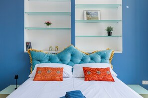 A bed with white bedding, two orange pillows, and a blue blanket. There are two glass shelves above the bed with a small plant and a picture frame.