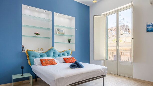 Une chambre à coucher avec un lit, des tables de chevet, une étagère en verre et une vue sur l’extérieur, visible à travers une porte.