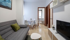 Una sala de estar con un sofá gris, un televisor, una mesa de comedor con sillas y una zona de cocina.