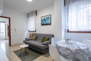 Uma sala de estar com um sofá cinza, uma mesinha branca e uma janela com cortinas leves.