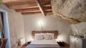 Una camera da letto con soffitto in legno, una grande parete in pietra, un letto con biancheria bianca, un comodino e un armadio.