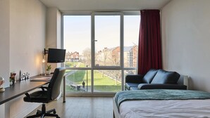 A hotel room with a large window, a desk with a computer, a chair, a bed, and a red curtain. - Upperton Road Studios in Leicester (Leicester)