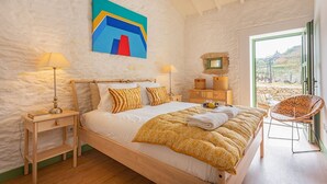 Un dormitorio con cama, mesitas de noche, una silla, un cuadro y vistas a través de una ventana.