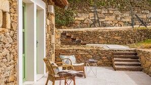 Un espacio al aire libre con paredes de piedra, mobiliario de mimbre y una pequeña mesa, junto a un edificio con una puerta verde.