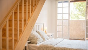 A bedroom with a wooden staircase, a bed with patterned bedding, a bedside table with a lamp, and a large window with a view of greenery outside.