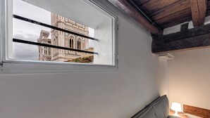 A room with a window overlooking a building, a bed with pillows, a nightstand with a lamp, and a wooden ceiling.
