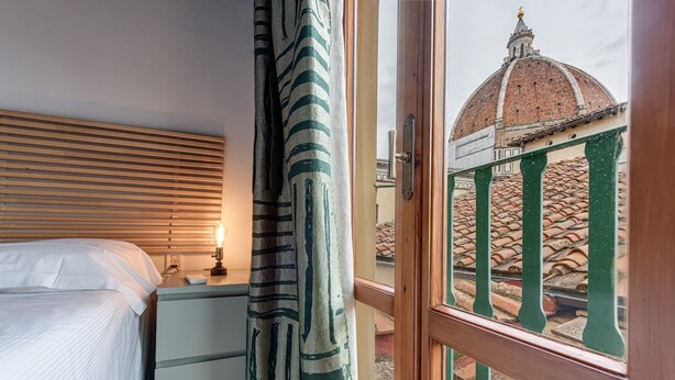 A bedroom with a bed, a nightstand, a lamp, and a view of a dome and tiled roof through a window.