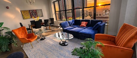 Una sala de estar moderna con un sofá azul, sillones naranjas, una mesa de centro de vidrio y ventanas grandes con vistas a la ciudad.