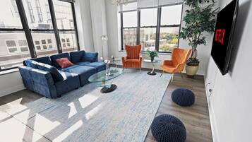 A modern living room with a sectional sofa, two armchairs, a glass coffee table, a large potted plant, and a flat-screen TV mounted on the wall.