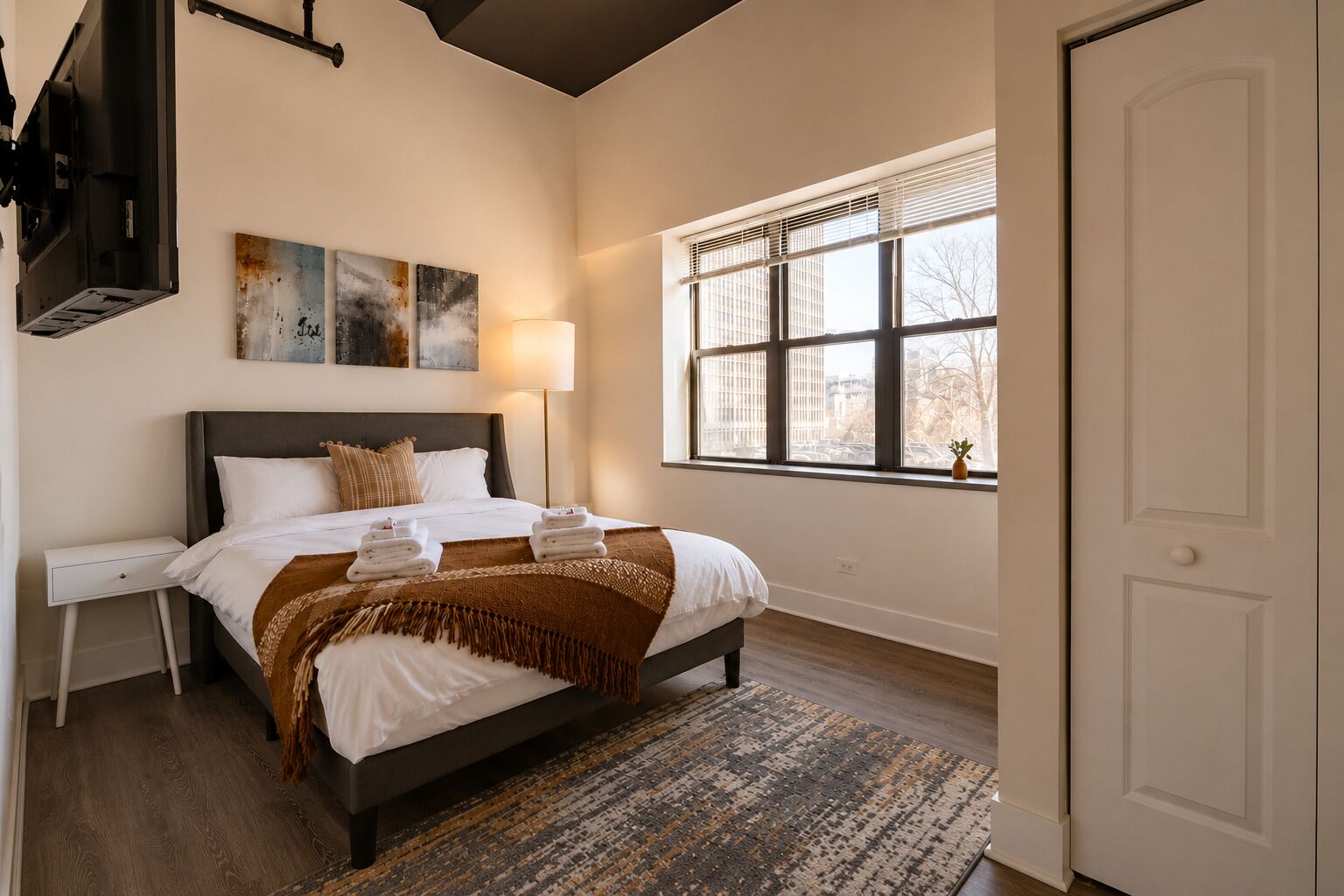 A bedroom with a bed, bedside tables, a TV, artwork, a window with blinds, and a door.