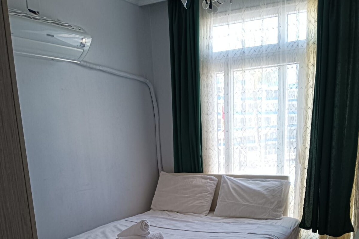 A single bed with white bedding and a rolled towel, a window with green curtains, and a tiled floor.