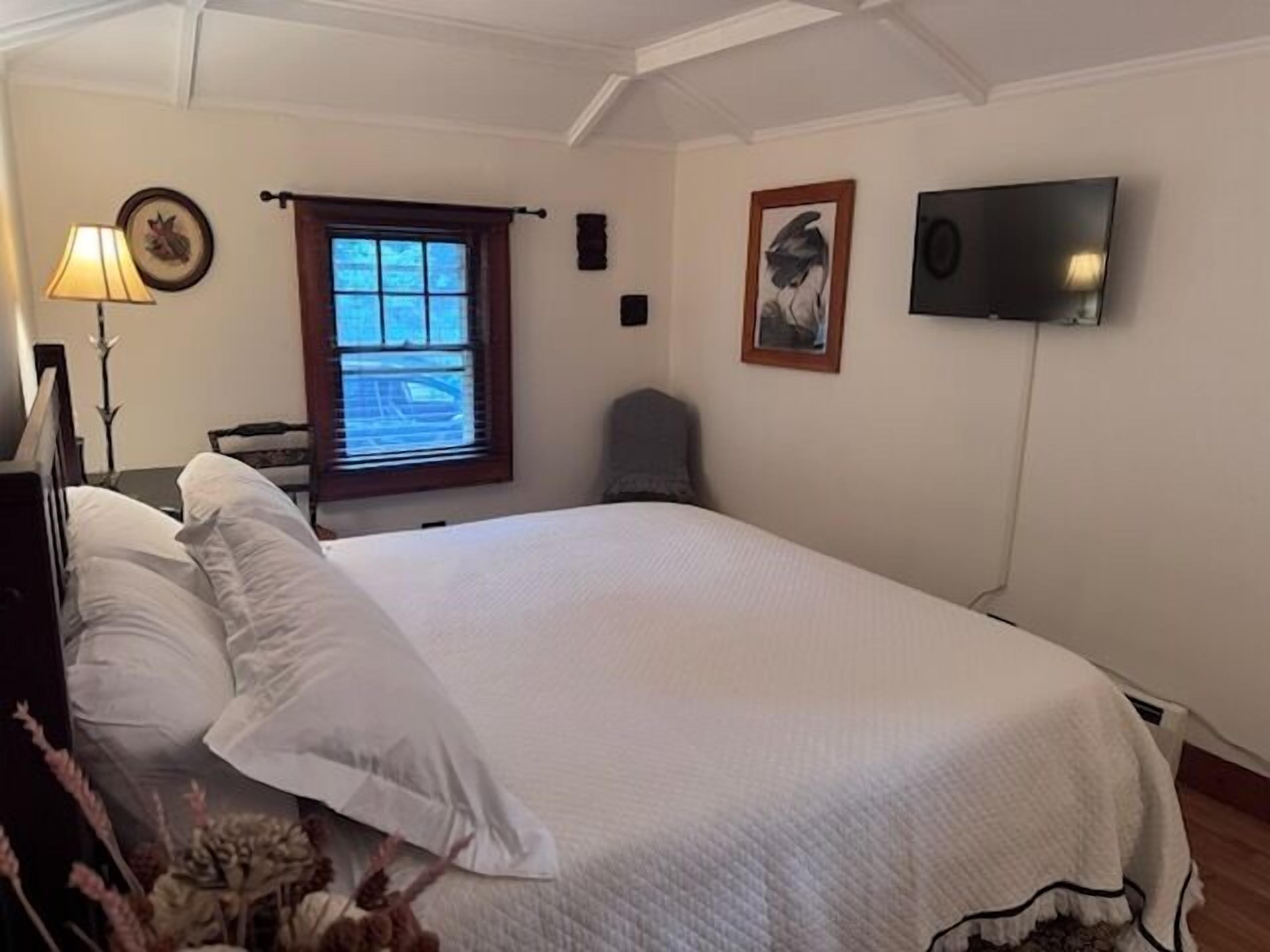 A bedroom with a large bed, a window with blinds, a lamp, and a flat-screen TV mounted on the wall.