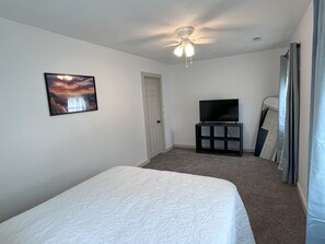 A hotel room with a bed, a television on a stand, a ceiling fan, a door, and a painting on the wall.